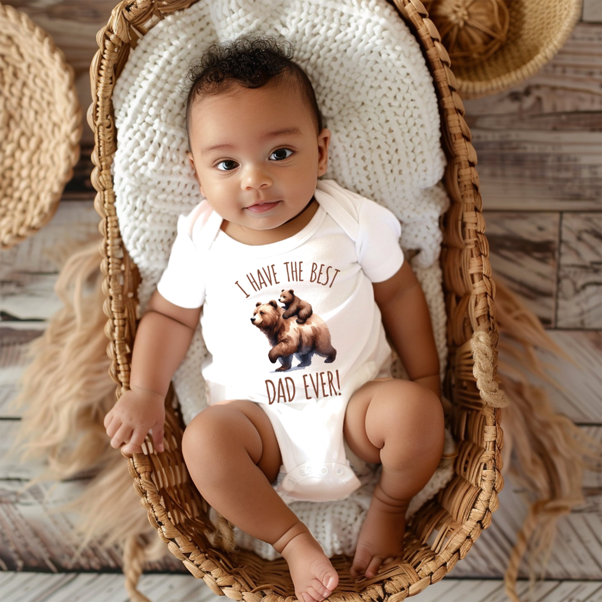 Trendy Tykes A baby with curly hair and big brown eyes sits in a woven basket on a textured blanket, wearing a white onesie with the text 'I have the BEST DAD EVER!' and a graphic of two bears above the text