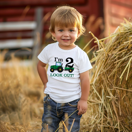 A toddler standing in a field with hay bales and a red barn in the background, wearing a white t-shirt with the text "I'm 2 LOOK OUT!" and a green tractor graphic, paired with denim jeans.