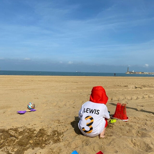 Trendy Tykes Small child named Lewis playing on a sandy beach, wearing a white t-shirt with 'LEWIS' and the number '3' in a black and white design