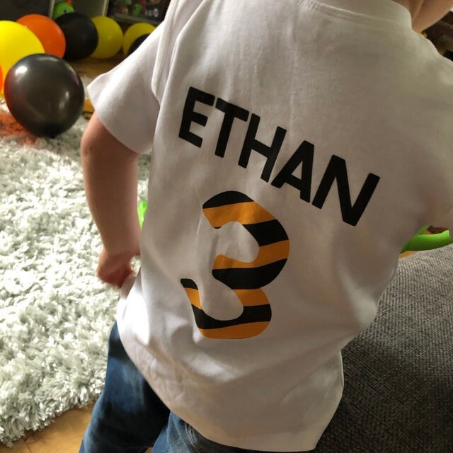 Trendy Tykes Back view of a young boy named Ethan, standing in a living room with balloons. He wears a white t-shirt personalized with 'ETHAN' and the number '3' in a black and orange striped design.