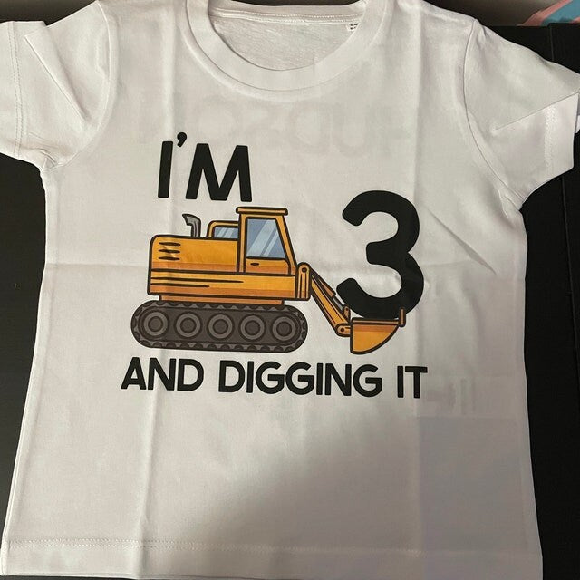 Trendy Tykes Flat lay of a white toddler t-shirt featuring a yellow bulldozer graphic with the phrase 'I'M 3 AND DIGGING IT' printed in black and orange.