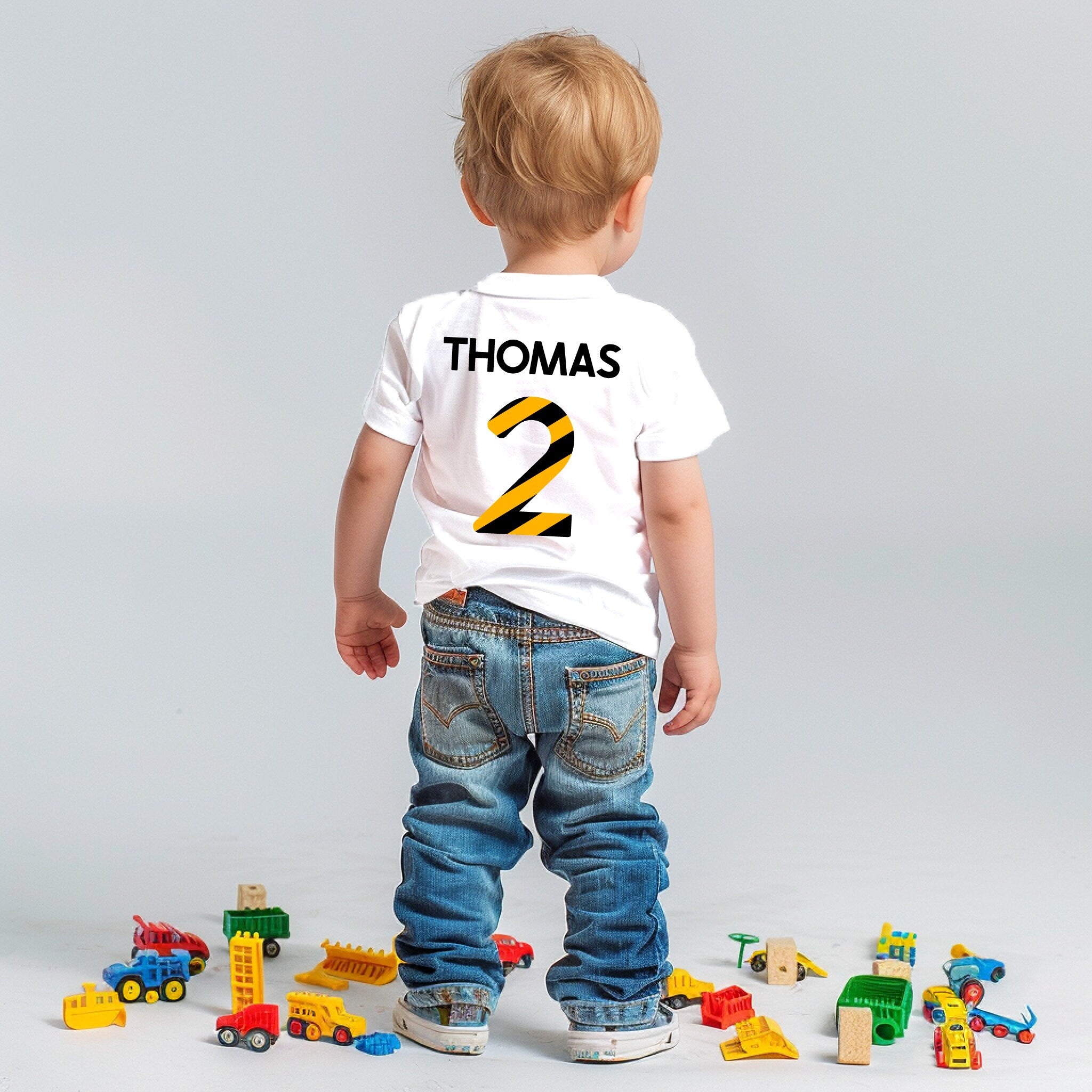 Back view of a young child in a white t-shirt with personalised back print with name and age in bold black and yellow construction tape pattern, standing amid scattered toys.