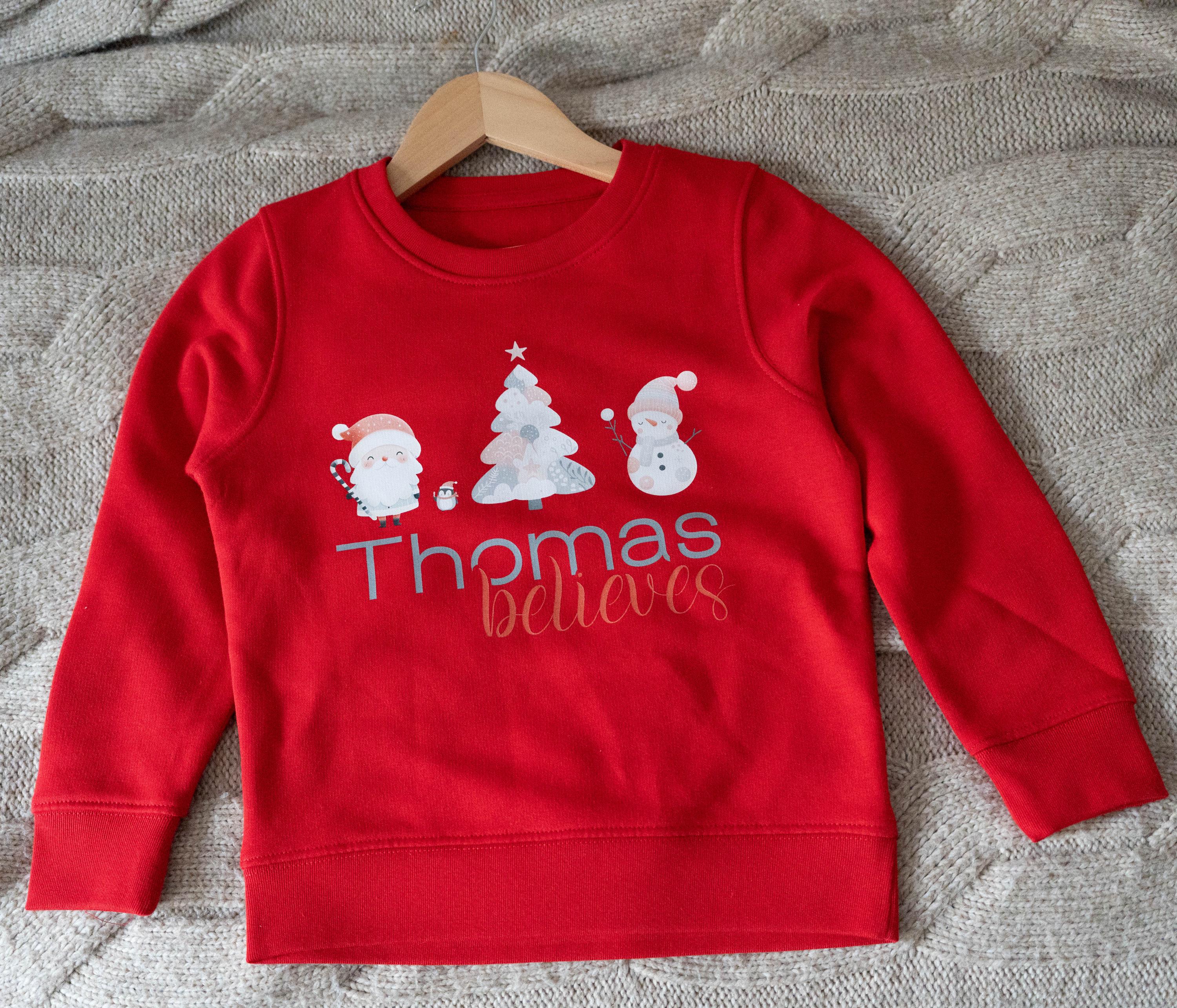 Red personalized children's Christmas jumper from Trendy Tykes featuring a festive design with Santa, a Christmas tree, a snowman, and the name 'Thomas' with the word 'believes' below, perfect for the holiday season.