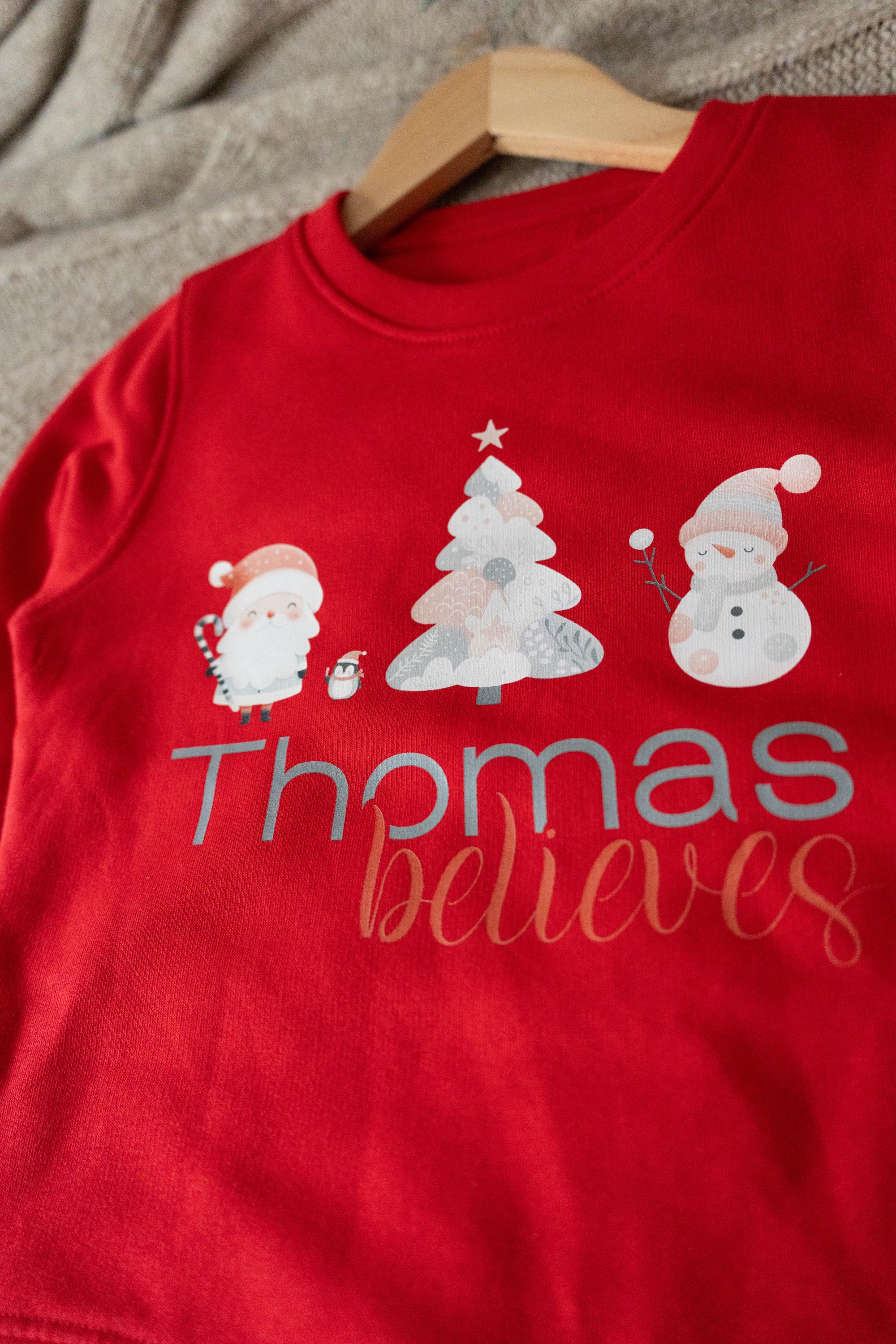 Red personalized children's Christmas jumper from Trendy Tykes featuring a festive design with Santa, a Christmas tree, a snowman, and the name 'Thomas' with the word 'believes' below, perfect for the holiday season.