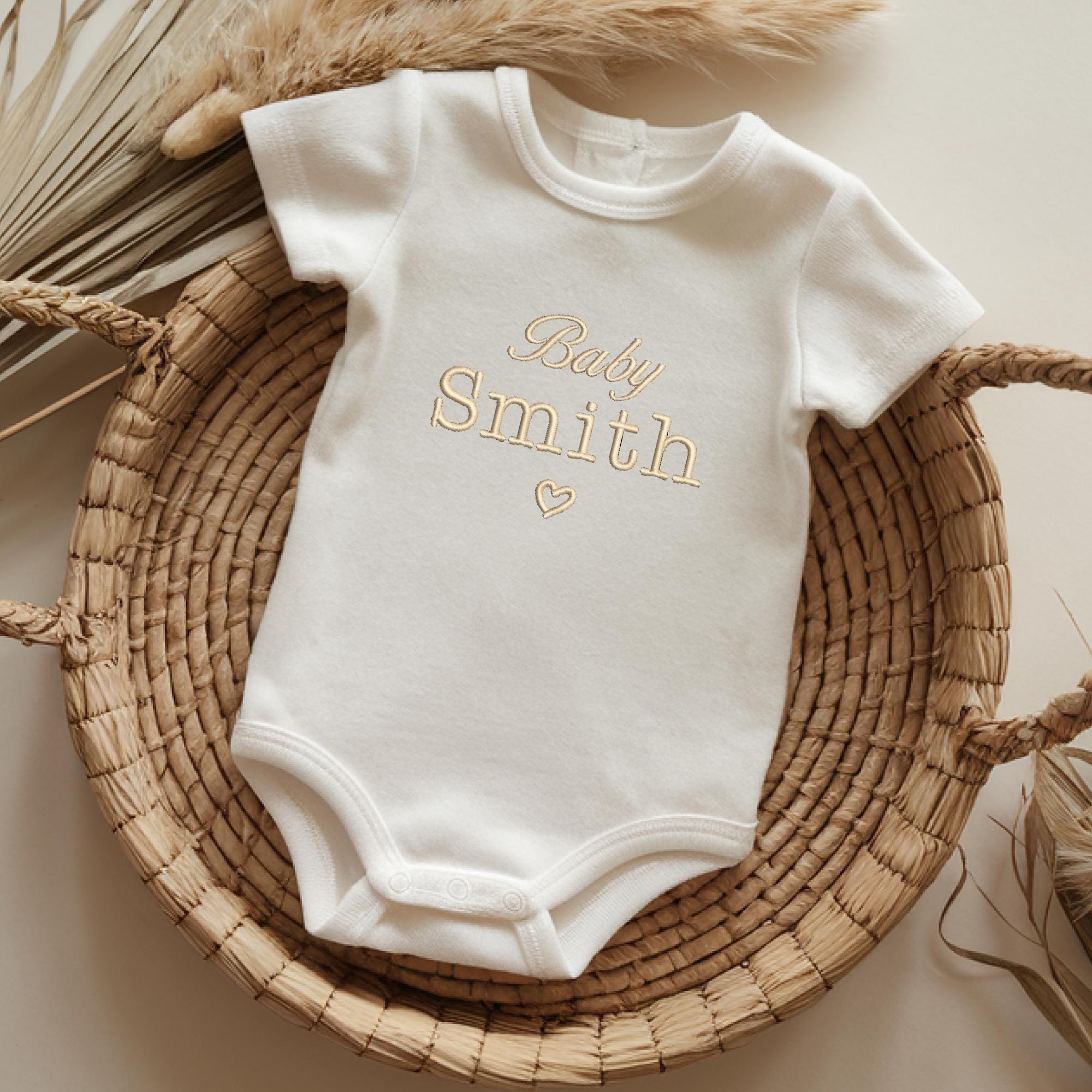 White baby bodysuit with embroidered text and a small heart underneath, displayed on a round woven basket with natural pampas grass accents for a warm and rustic look.