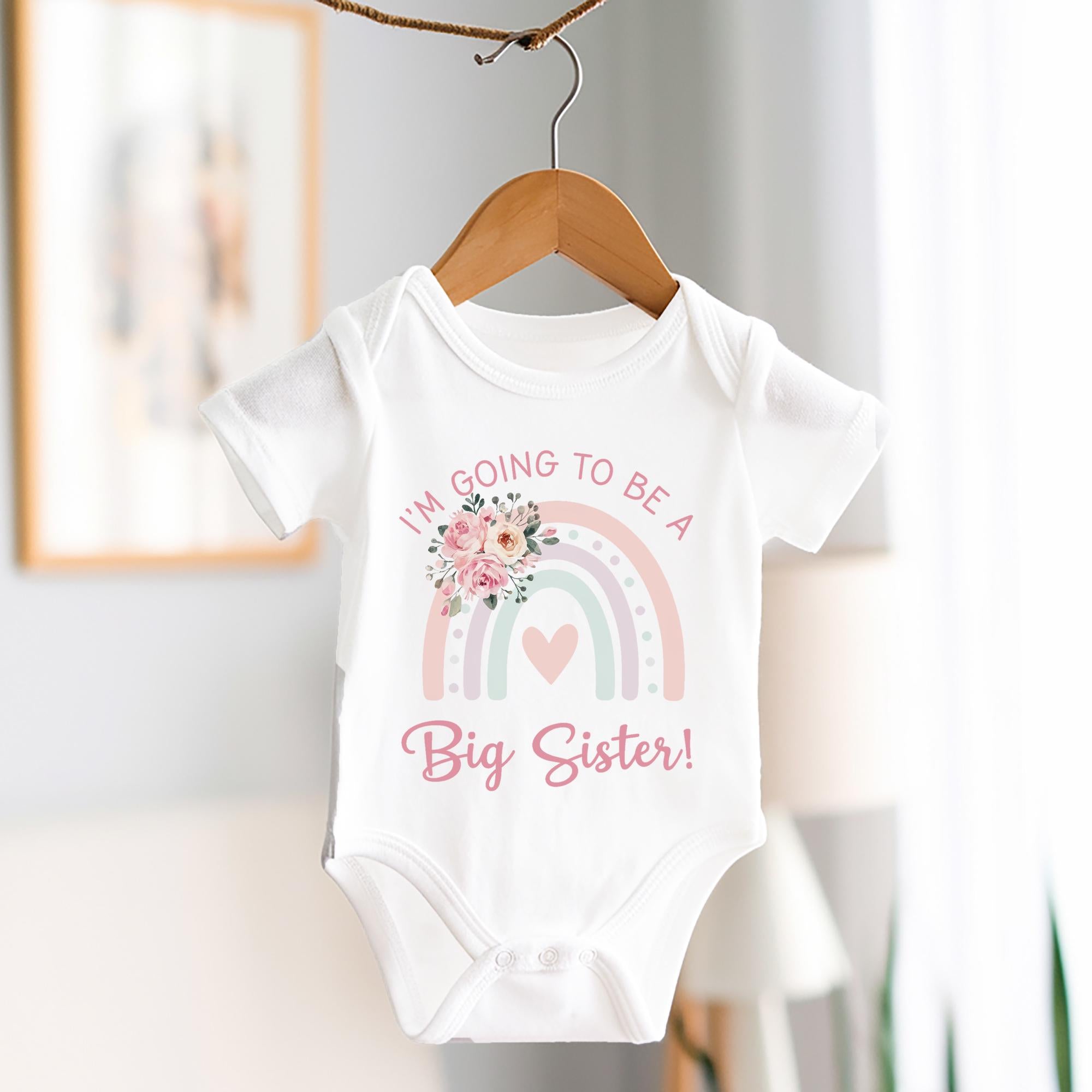 A white baby vest on a wooden hanger featuring a pastel rainbow design with a heart in the center. The text reads "I'M GOING TO BE A Big Sister!" in pink and pastel colors. A floral graphic with pink roses decorates the top left of the rainbow