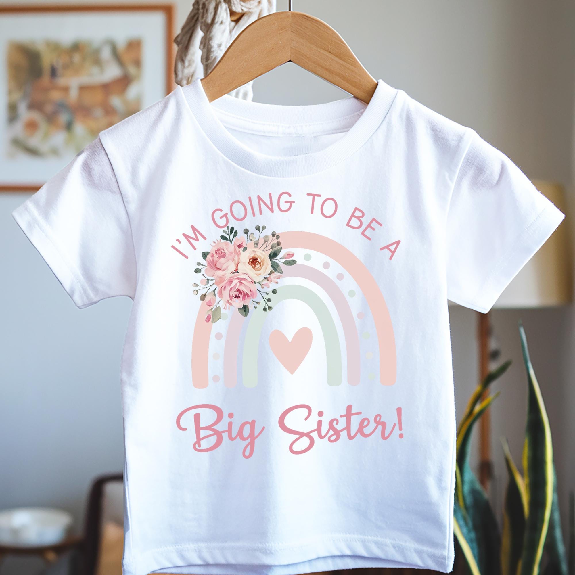 A white t-shirt on a wooden hanger featuring a pastel rainbow design with a heart in the center. The text reads "I'M GOING TO BE A Big Sister!" in pink and pastel colors. A floral graphic with pink roses decorates the top left of the rainbow.
