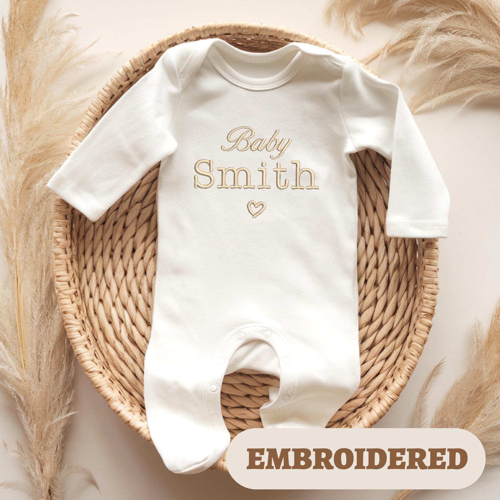 White baby romper with the embroidered text and a small heart beneath, displayed on a woven basket surrounded by soft pampas grass for a cozy and neutral aesthetic. A label in the corner reads 'EMBROIDERED' in bold text.