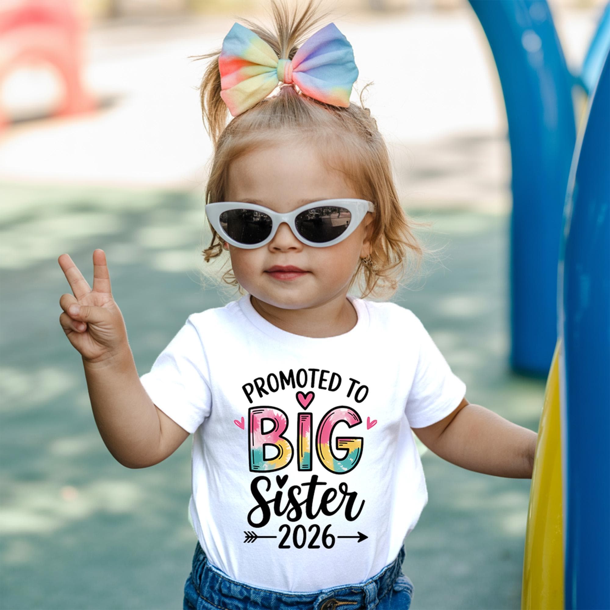 Smiling toddler girl wearing sunglasses and a rainbow bow, posing in a white T-shirt that reads “Promoted to Big Sister 2026” – perfect for sibling announcement or pregnancy reveal.