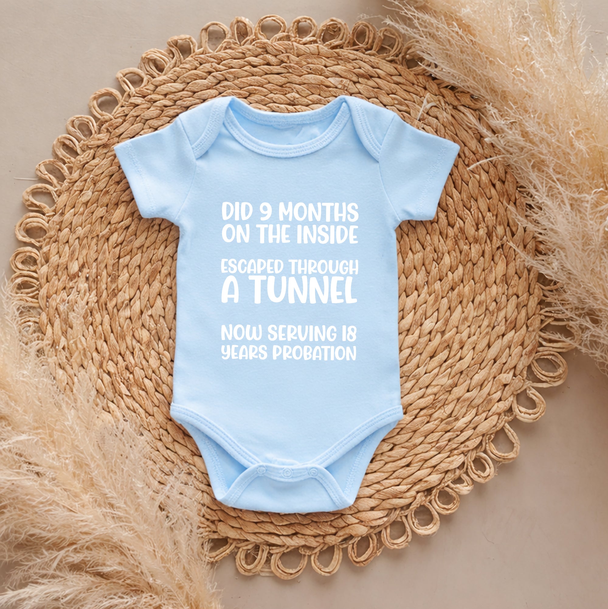 Light blue baby onesie with humorous text on a woven mat