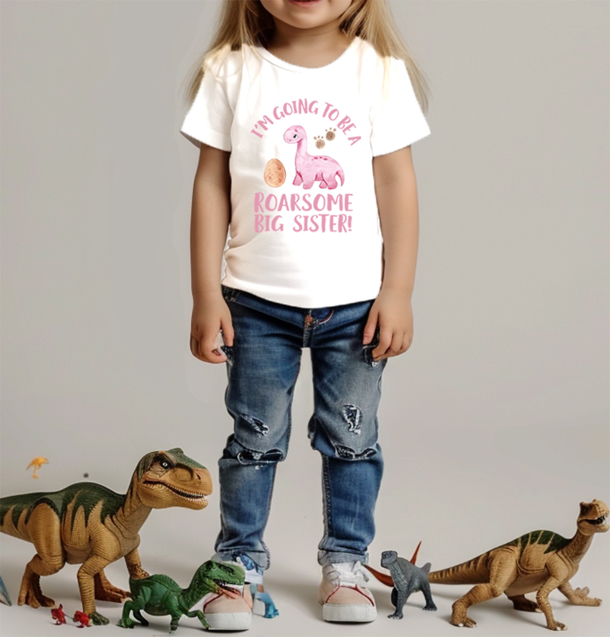 A young child stands with hands in jean pockets, wearing a Trendy Tykes white T-shirt with a "roarsome big sister" dinosaur graphic, surrounded by toy dinosaurs.