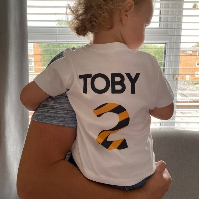 Trendy Tykes Small child named Toby, being held, showing the back of his white t-shirt personalized with &#39;TOBY&#39; and the number &#39;2&#39; in black and orange striped design,
