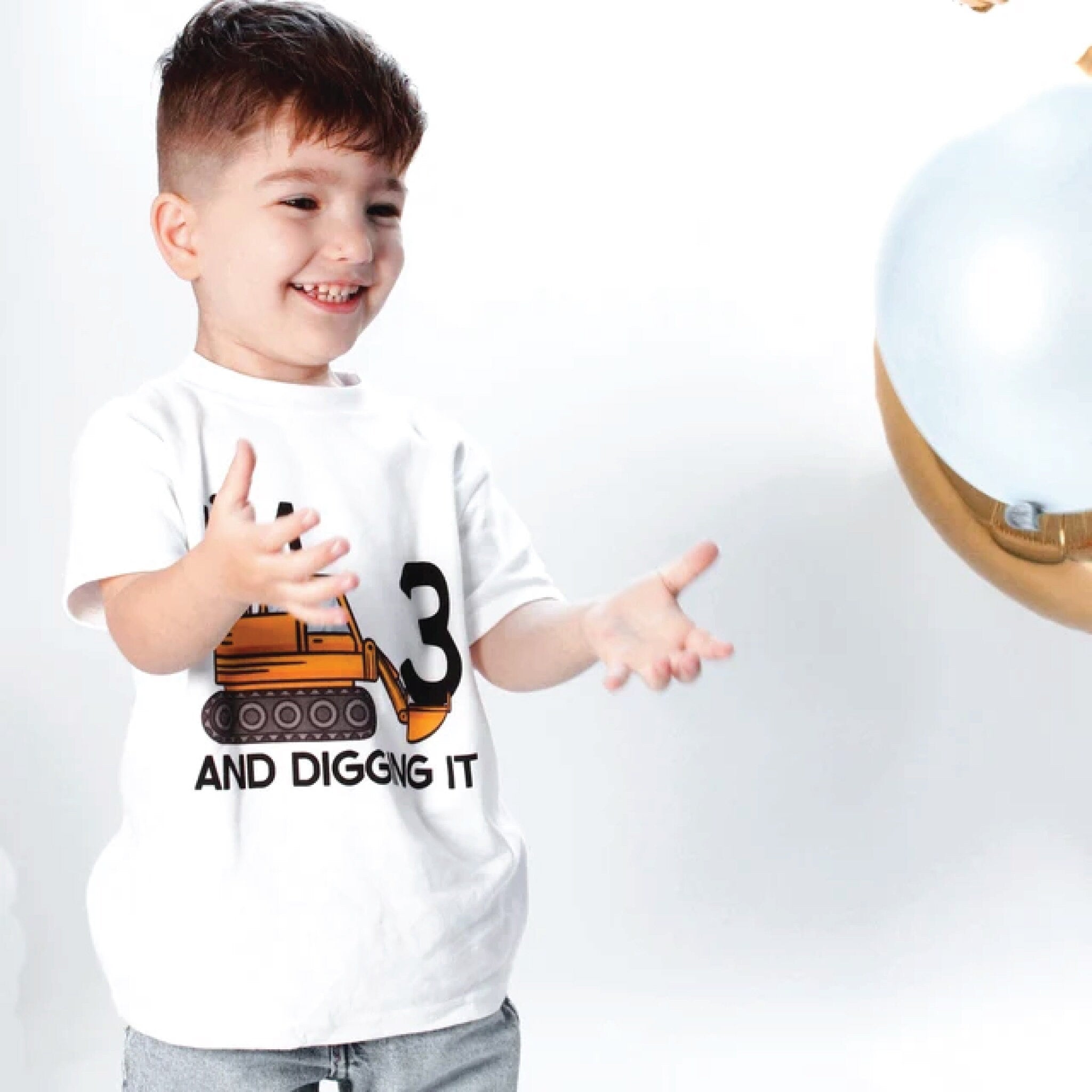 Trendy Tykes Cheerful young boy with short brown hair playing with a large gold balloon, wearing a white t-shirt with a graphic of a yellow bulldozer and the text &#39;3 AND DIGGING IT&#39; to celebrate his third birthday.