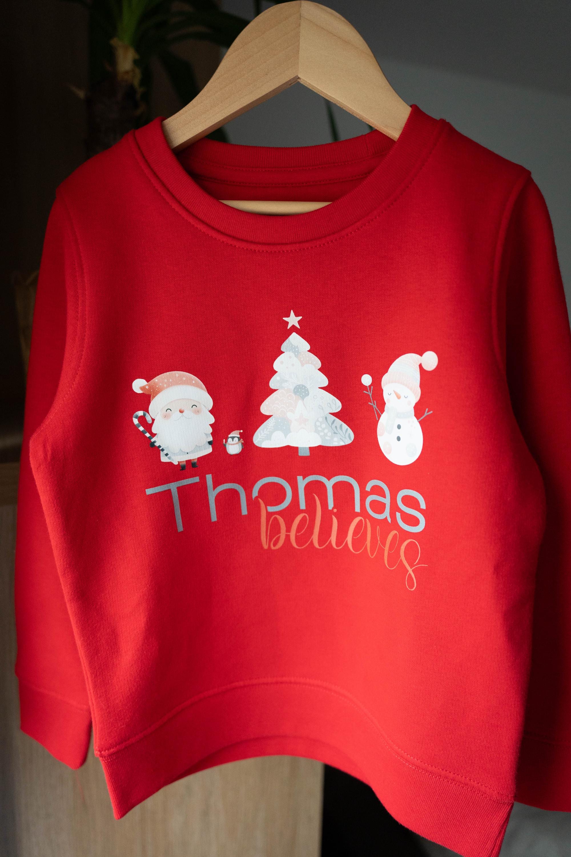 Red personalized children&#39;s Christmas jumper from Trendy Tykes featuring a festive design with Santa, a Christmas tree, a snowman, and the name &#39;Thomas&#39; with the word &#39;believes&#39; below, perfect for the holiday season.