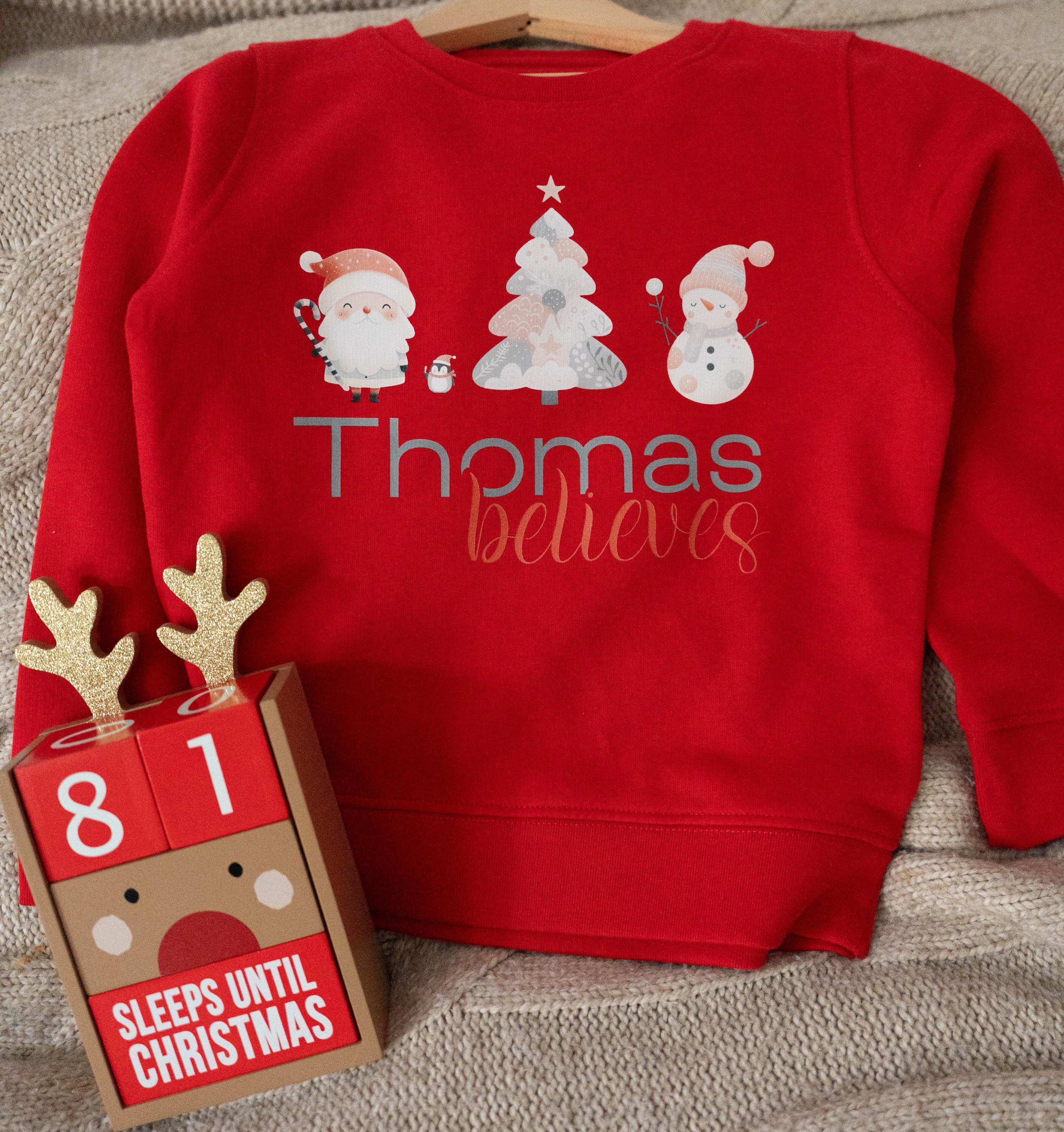 Red personalized children&#39;s Christmas jumper from Trendy Tykes featuring a festive design with Santa, a Christmas tree, a snowman, and the name &#39;Thomas&#39; with the word &#39;believes&#39; below, perfect for the holiday season.