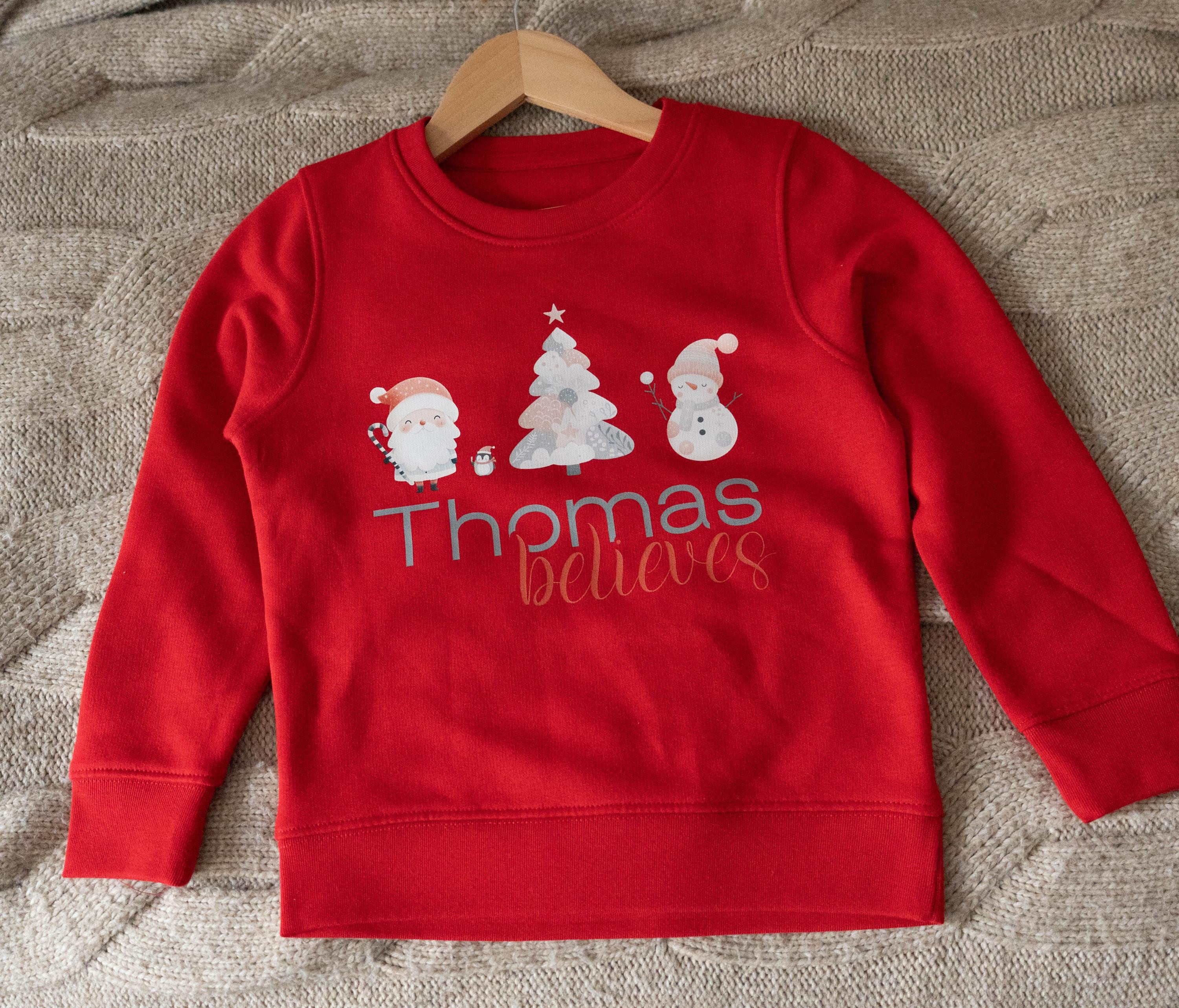 Red personalized children&#39;s Christmas jumper from Trendy Tykes featuring a festive design with Santa, a Christmas tree, a snowman, and the name &#39;Thomas&#39; with the word &#39;believes&#39; below, perfect for the holiday season.