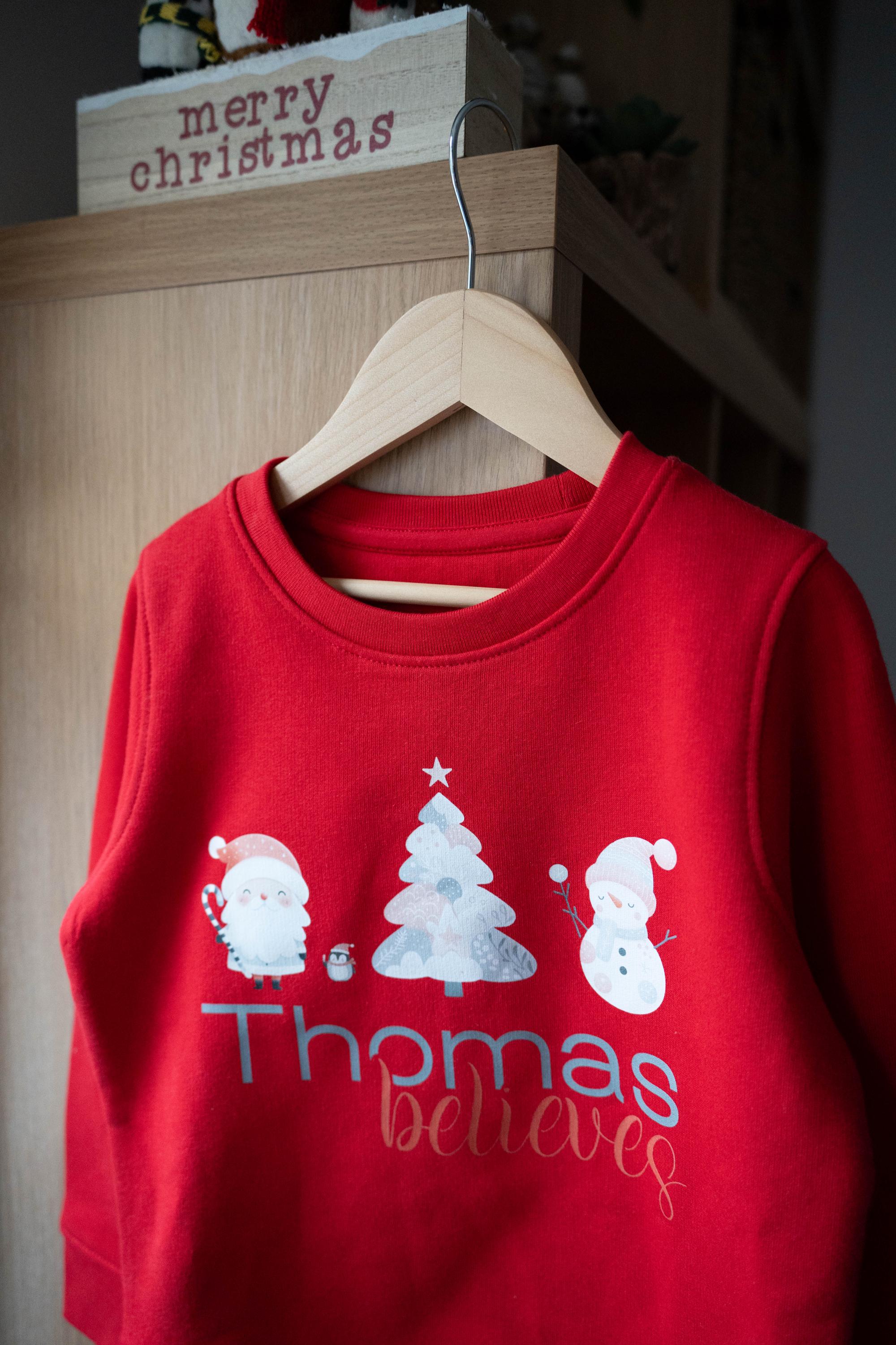 Red personalized children&#39;s Christmas jumper from Trendy Tykes featuring a festive design with Santa, a Christmas tree, a snowman, and the name &#39;Thomas&#39; with the word &#39;believes&#39; below, perfect for the holiday season.