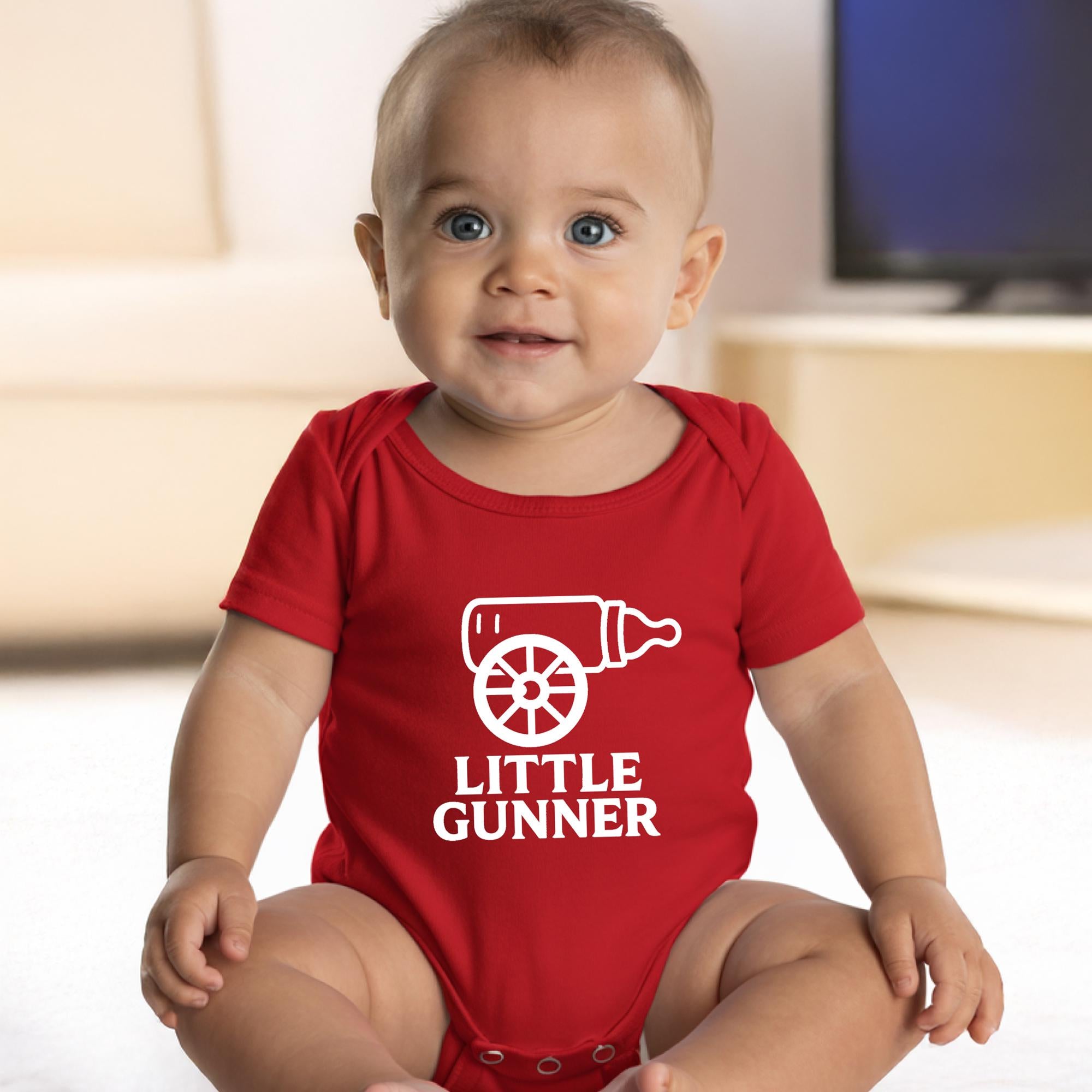 Smiling baby wearing a red Little Gunner baby vest with white milk bottle cannon design, sitting indoors – fun football fan baby outfit and gift idea.