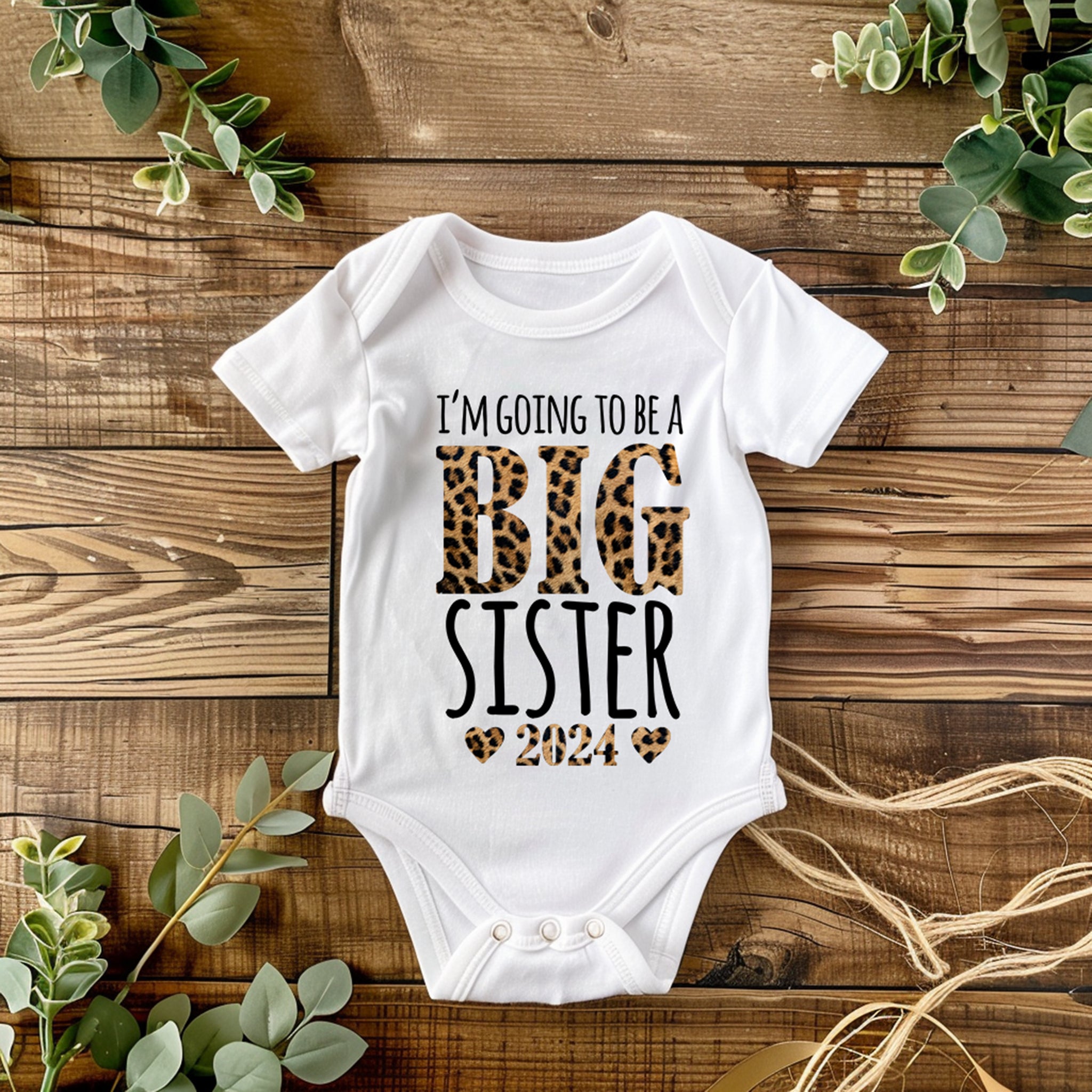 Trendy Tykes a baby bodysuit with the phrase "I'm going to be a Big Sister 2024" printed in a playful mix of bold and leopard print fonts. The design includes cute heart-shaped accents, adding a charming and celebratory touch. 