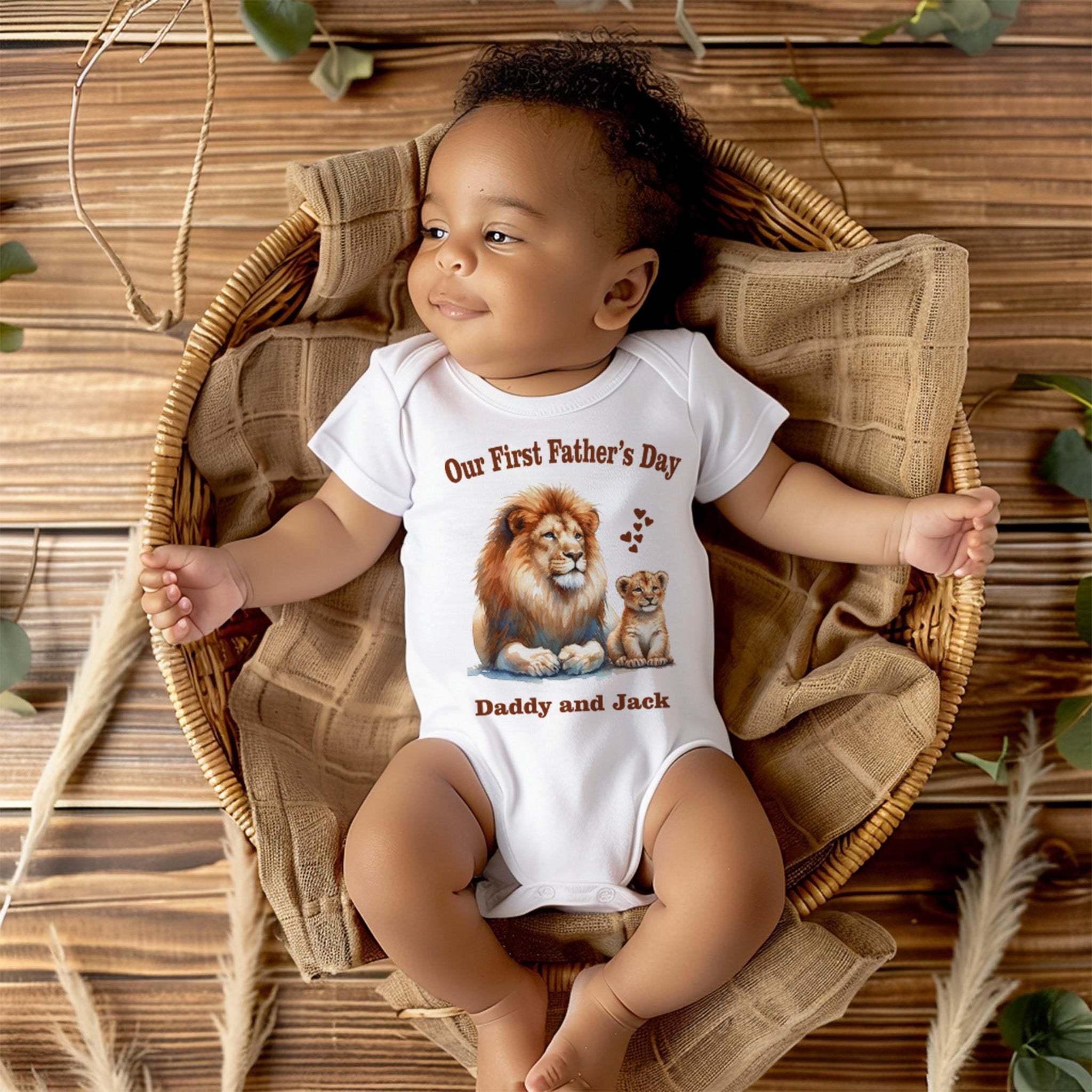 Trendy Tykes A cheerful baby with curly hair lies contentedly in a woven basket, wearing a white bodysuit adorned with the text 'Our First Father's Day' in a warm, inviting font. Below the text is an affectionate illustration of a lion and a cub, symbolizing the bond between father and child. The names 'Daddy and Jack' are personalized underneath the illustration, making this bodysuit a special keepsake for their first Father's Day celebration together.